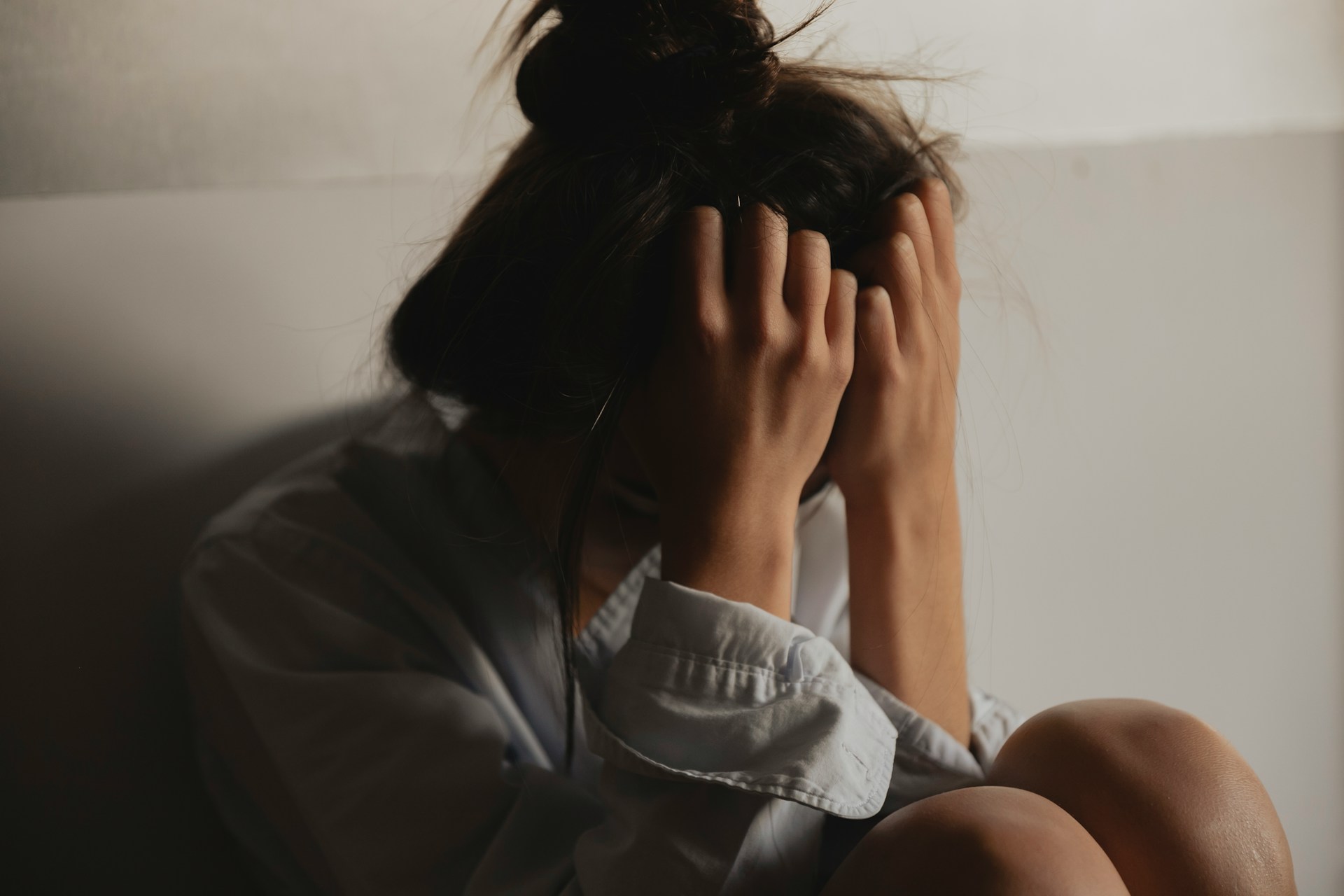A person sitting against a plain wall with their head in their hands, wearing a light-colored button-up shirt. The photo is taken in dim, moody lighting.